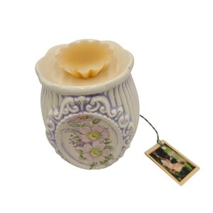 May include: A ceramic wax warmer with a floral design. The warmer is light purple with a cream-colored top and a floral wax melt. The warmer has a decorative floral pattern and a small tag attached.