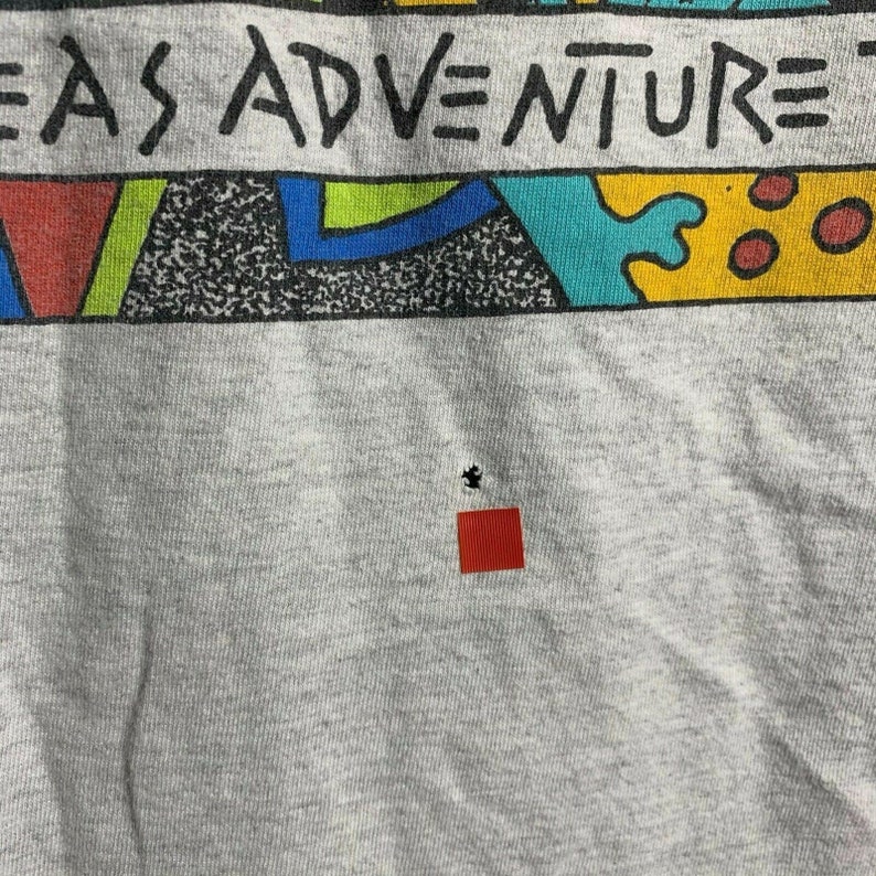 Pode incluir: Camiseta cinza com um design geom&eacute;trico colorido e o texto "Ideas Adventure". A camiseta tem um pequeno furo no centro.