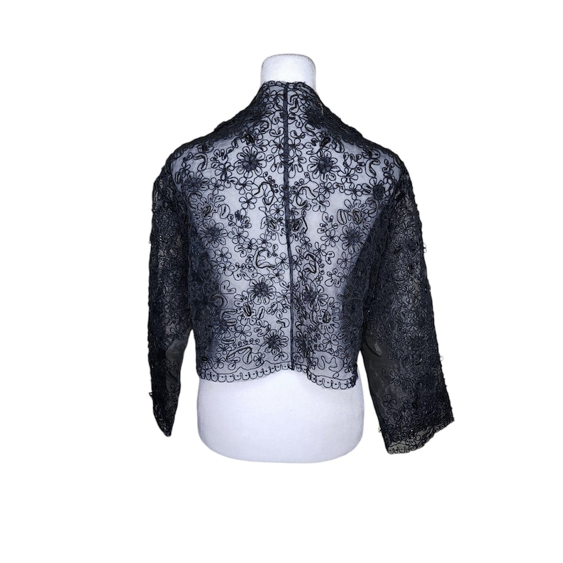 May include: A black lace shrug with a floral pattern. The shrug is cropped and has three-quarter sleeves.