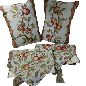 Op de afbeelding: Twee decoratieve kussens en twee tafellopers. De kussens en lopers hebben een lichtblauwe achtergrond met een herhalend patroon van rode appels, groene bladeren en bruine takken. De kussens hebben een geschulpte rand met een rood-groen ruitpatroon.