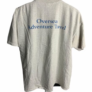 Pode incluir: Uma camiseta cinza com o texto "Overseas Adventure Travel" impresso em azul nas costas.