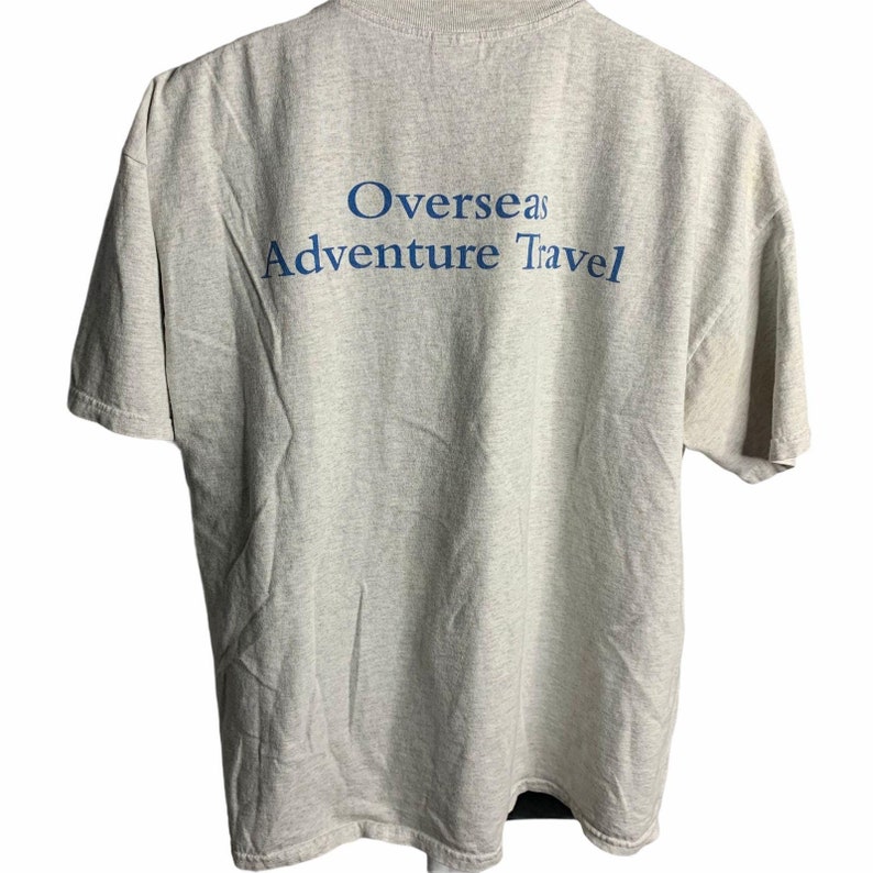 Pode incluir: Uma camiseta cinza com o texto "Overseas Adventure Travel" impresso em azul nas costas.