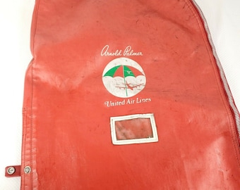 Pode incluir: Bolsa de roupa vermelha com um zíper branco e uma etiqueta branca. A bolsa tem um logotipo de guarda-chuva verde e branco com o texto "Arnold Palmer" e "United Air Lines".