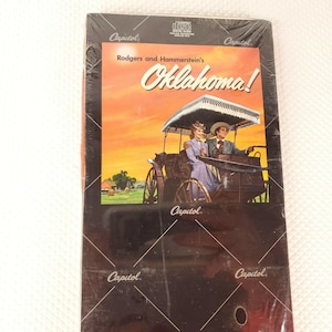 Puede incluir: Una imagen en blanco y negro de una portada de álbum de disco vintage con una pareja montando en un carruaje tirado por caballos. El álbum se titula "Oklahoma!" de Rodgers y Hammerstein. El álbum está en el sello Capitol Records.