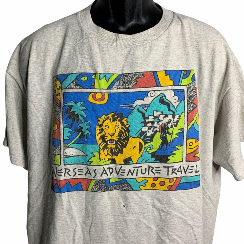 Pode incluir: Uma camiseta cinza com uma estampa gr&aacute;fica colorida de um le&atilde;o, montanhas e uma pessoa fazendo caminhadas. O gr&aacute;fico &eacute; cercado por uma borda colorida com o texto "Overseas Adventure Travel".