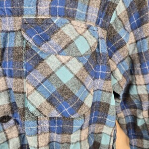 Puede incluir: Primer plano de una camisa de franela a cuadros azules, grises y turquesas. La camisa tiene un bolsillo en el pecho y cierre de botones. La tela es suave y parece ser de algod&oacute;n o similar. Es un estilo casual.