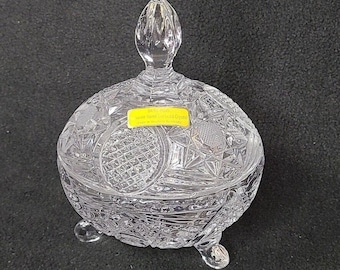 May include: A clear, hand-cut crystal candy dish with a lid and three small feet. The dish features intricate geometric patterns and a teardrop-shaped handle on the lid. A yellow sticker with text is visible on the dish.