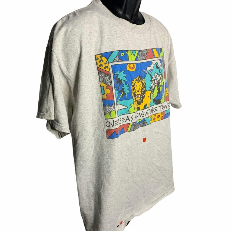 Pode incluir: Uma camiseta cinza com uma estampa gr&aacute;fica colorida de um le&atilde;o em uma cena de selva. O gr&aacute;fico inclui o texto "Overseas Adventure Travel".