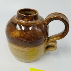 Może przedstawiać: Ręcznie wykonany ceramiczny kubek o zaokrąglonym korpusie i dużym, pętelkowatym uchwycie. Kubek ma dwutonowe szkliwo, z jasnobeżową podstawą i bogatą brązową górną częścią. Uchwyt jest również brązowy.