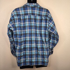 Puede incluir: Una camisa de franela a cuadros azules, grises y turquesas. La camisa tiene mangas largas y una parte delantera abotonada. El estampado de cuadros es un dise&ntilde;o cl&aacute;sico, adecuado para ropa informal. La camisa se muestra en una percha negra.