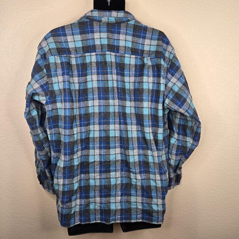 Puede incluir: Una camisa de franela a cuadros azules, grises y turquesas. La camisa tiene mangas largas y una parte delantera abotonada. El estampado de cuadros es un dise&ntilde;o cl&aacute;sico, adecuado para ropa informal. La camisa se muestra en una percha negra.