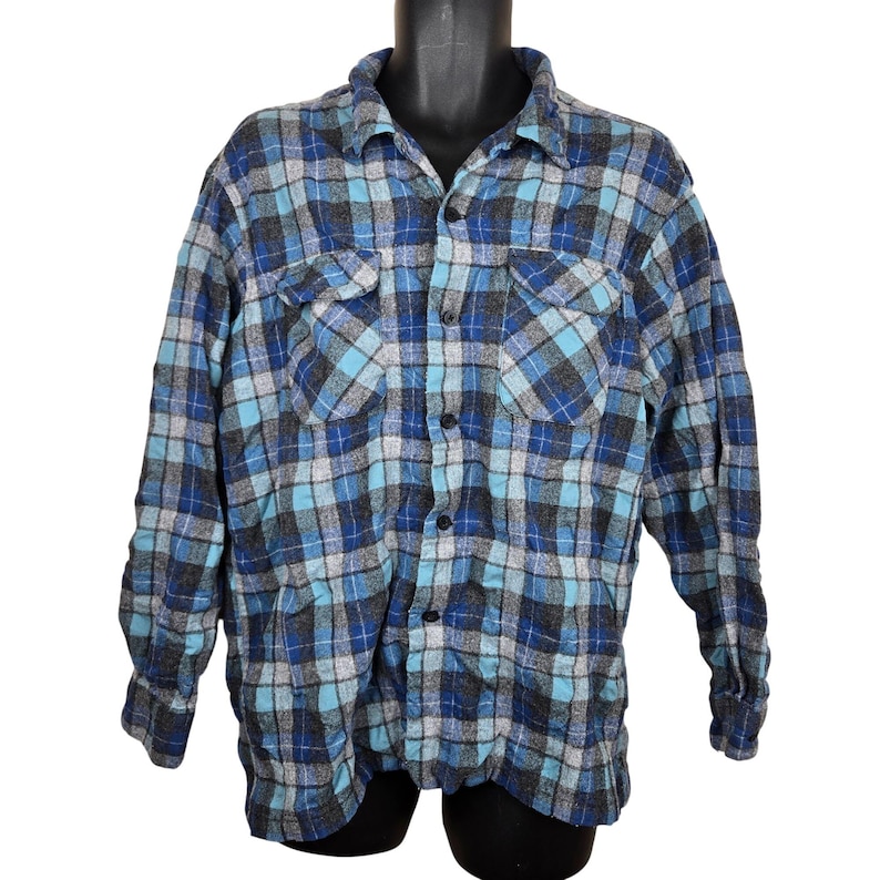 Puede incluir: Camisa de franela a cuadros azules, grises y turquesas con dos bolsillos en el pecho. La camisa de manga larga tiene una botonadura frontal y un cuello cl&aacute;sico. La tela parece suave y c&oacute;moda.