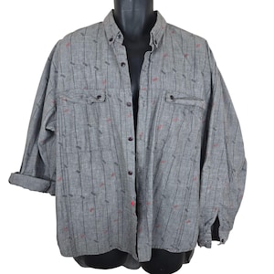Pode incluir: Camisa cinza com botões, com listras verticais pretas e pequenas formas geométricas vermelhas e cinzas. A camisa de manga comprida tem dois bolsos no peito e as mangas estão enroladas. A camisa está abotoada.
