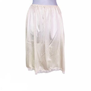 May include: Cream-colored half slip with a textured fabric and a delicate lace trim at the hem. The slip has a gathered waistband and a flowing, A-line silhouette. This vintage undergarment is designed to be worn under skirts.
