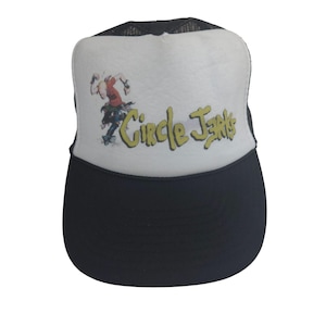 May include: A white and black trucker hat with a black mesh back. The front of the hat features a cartoon illustration of a person skateboarding with the text "Circle Jerks" in yellow.