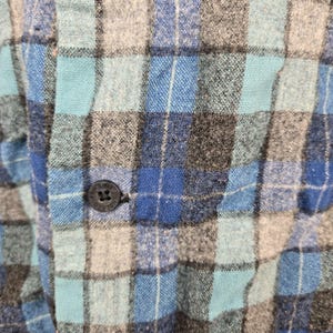 Puede incluir: Primer plano de una camisa de franela a cuadros azules, grises y turquesas. La camisa tiene un cierre de botones con un solo bot&oacute;n visible. La tela tiene un aspecto texturizado.