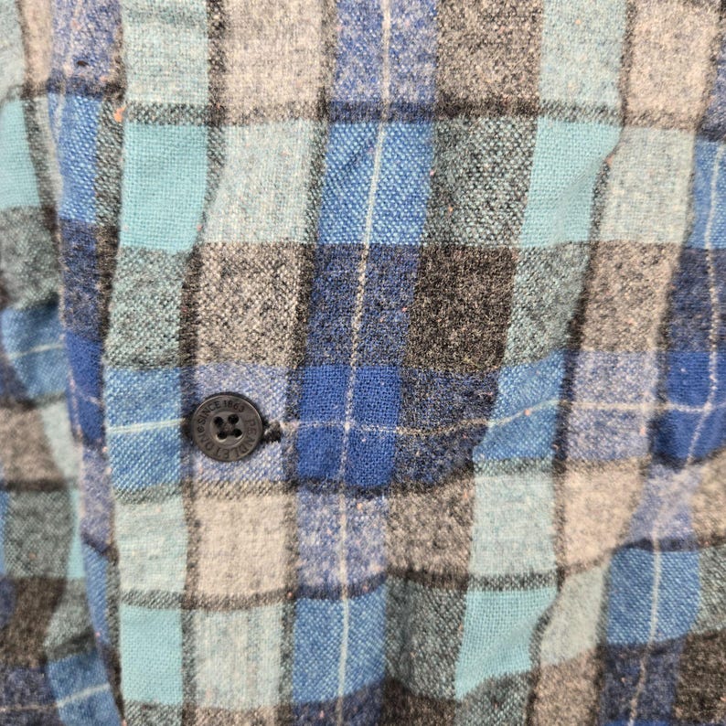 Puede incluir: Primer plano de una camisa de franela a cuadros azules, grises y turquesas. La camisa tiene un cierre de botones con un solo bot&oacute;n visible. La tela tiene un aspecto texturizado.