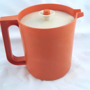 May include: Orange plastic pitcher with a white lid and handle. The pitcher has a rounded body and a spout. The lid has a small orange knob. The handle is a curved, open design. The pitcher is likely vintage.