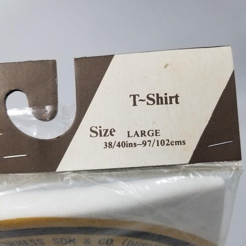May include: A white T-shirt, size large, is visible through a clear plastic package. The label reads "T-Shirt" and includes size details: 38/40 inches, equivalent to 97/102 centimeters. The packaging has a brown and white design.