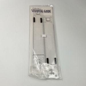 May include: A clear plastic stamping guide in a clear plastic package. The guide has two long rulers with measurement markings and the text "ColorBox STAMPING GUIDE" at the top. The guide is designed for crafting and stamping projects.