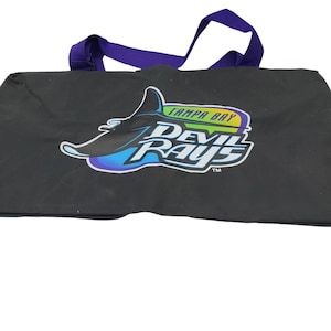 Puede incluir: Bolso tote negro con asas moradas con el logotipo de los Tampa Bay Devil Rays. El logotipo incluye una manta raya estilizada en blanco y azul, con el nombre del equipo en verde y azul.