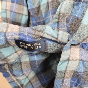 Puede incluir: Primer plano de una camisa a cuadros azul, gris y blanca. La camisa tiene una etiqueta que dice "1961 ORIGINAL SURF PLAID". La tela es suave y parece franela.