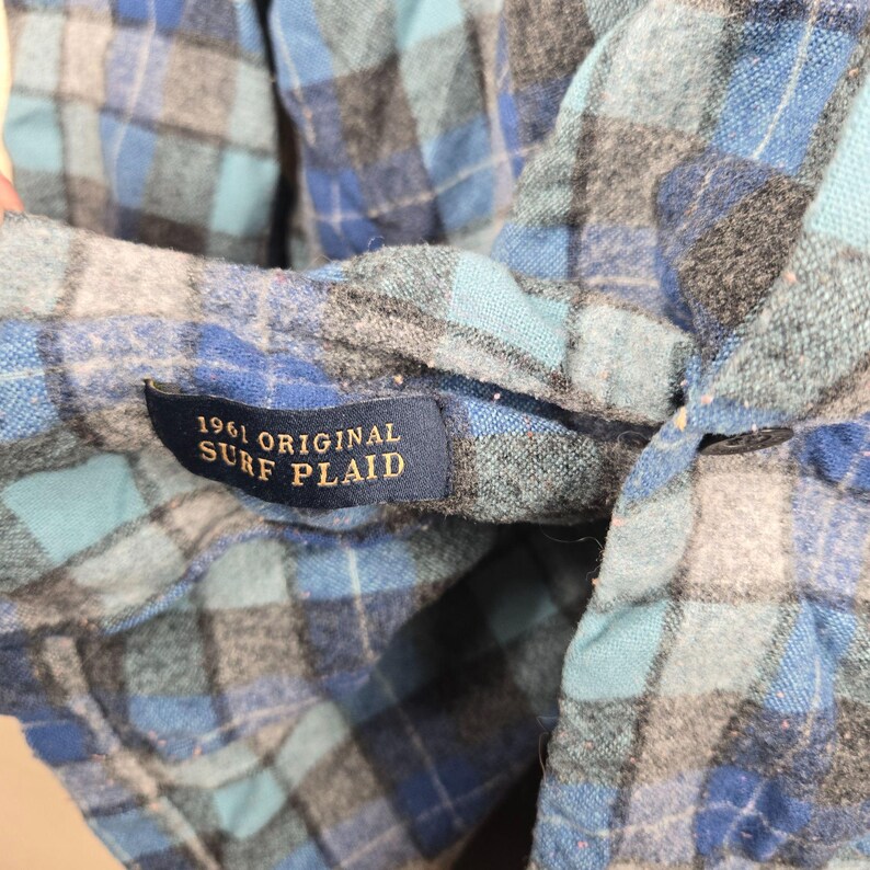 Puede incluir: Primer plano de una camisa a cuadros azul, gris y blanca. La camisa tiene una etiqueta que dice "1961 ORIGINAL SURF PLAID". La tela es suave y parece franela.