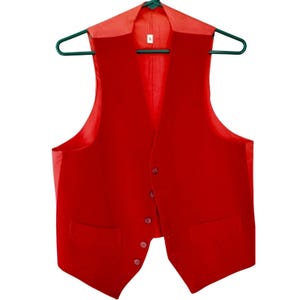 May include: A vibrant red waistcoat with a traditional design, featuring a notched lapel, two front pockets, and a button-up closure. The waistcoat is suspended on a green hanger. The garment appears to be made of a smooth, possibly velvet-like material.
