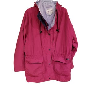 May include: A magenta-colored hooded jacket with a drawstring hood and waist. The jacket has two front pockets, a button-up closure, and the Woolrich logo on the chest. The interior lining is light gray.