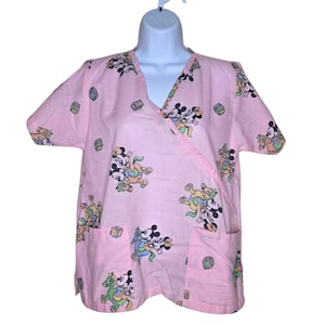 May include: Pink wrap-style scrub top with a V-neck and short sleeves. The top features a pattern of Mickey and Minnie Mouse riding carousel horses and blocks. Two pockets are visible on the front.