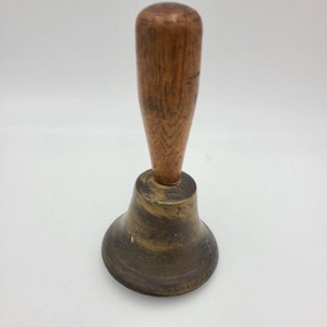 Puede incluir: Una campana de latón con un mango de madera. La campana tiene una base acampanada y un acabado ligeramente deslustrado. El mango es de color marrón claro y parece ser de madera. La campana probablemente se utiliza para señalización o decoración. Altura aproximada 15 cm.