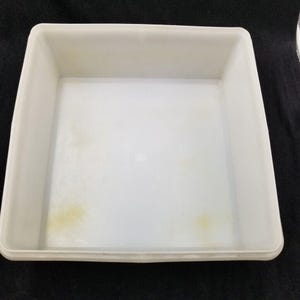 May include: A white, square plastic tray with slightly rounded corners. The tray has a shallow depth and appears to be empty. Some yellow discoloration is visible on the interior surface. The tray is set against a black background.