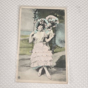 Può includere: Due donne in abiti vintage e grandi cappelli colorati sono sedute su un'altalena. La donna in primo piano indossa un vestito rosa con balze e un cappello blu e bianco con fiori. La donna dietro di lei indossa un vestito blu e un cappello bianco con fiori.