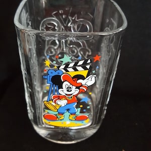 Puede incluir: Vaso de vidrio transparente con un diseño de Mickey Mouse con temática de cine. Mickey lleva un sombrero rojo y pantalones azules, sosteniendo un megáfono. El vaso tiene forma cuadrada y está decorado con estrellas y una claqueta.