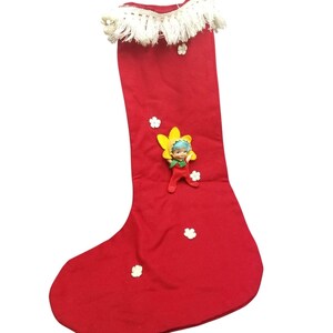 May include: A red Christmas stocking with a white fringe trim at the top. The stocking is decorated with small white flower-shaped embellishments and a small doll figure with a yellow flower. The doll is wearing a red outfit.