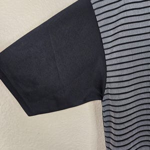 May include: Black short-sleeved shirt with a contrasting design. The left sleeve is solid black, while the right side features a pattern of thin, vertical white stripes on a black background. The fabric appears to be a lightweight material.