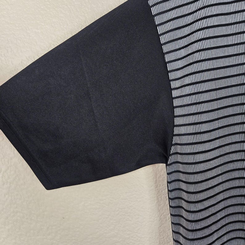 May include: Black short-sleeved shirt with a contrasting design. The left sleeve is solid black, while the right side features a pattern of thin, vertical white stripes on a black background. The fabric appears to be a lightweight material.