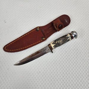 May include: A vintage hunting knife with a brown leather sheath. The knife features a stainless steel blade, a handle with a bone-like grip, and a metal pommel. The sheath is a reddish-brown color with a snap closure.