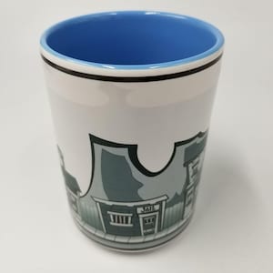 May include: White ceramic mug with a blue interior and a black rim. The mug features a cartoon illustration of a western town, including a building labeled "JAIL". The mug is cylindrical and appears to be unused.