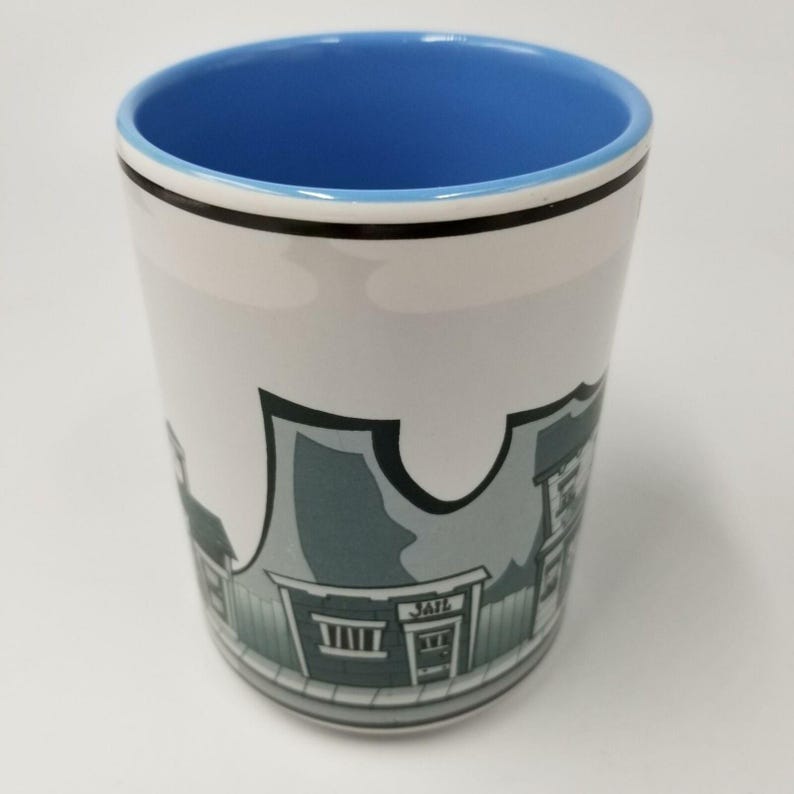 May include: White ceramic mug with a blue interior and a black rim. The mug features a cartoon illustration of a western town, including a building labeled "JAIL". The mug is cylindrical and appears to be unused.