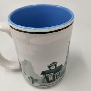 May include: White ceramic mug with a blue interior and a handle. The mug features a black rim and a cartoon illustration of a desert scene with a building and cactus. The word "Scenic" is visible on the building.