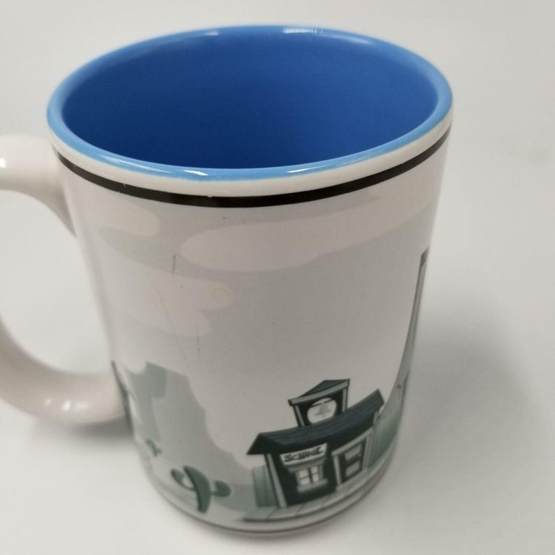 May include: White ceramic mug with a blue interior and a handle. The mug features a black rim and a cartoon illustration of a desert scene with a building and cactus. The word "Scenic" is visible on the building.