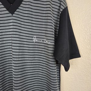 May include: A black and gray striped V-neck shirt with short sleeves. The shirt has a solid black sleeve and a white signature on the front. The shirt is made of a soft, comfortable material.