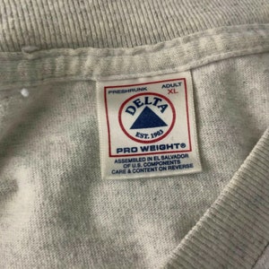 Pode incluir: Uma etiqueta branca com texto preto que diz "Preshrunk Adult XL Delta Est. 1903 Pro Weight Assembled in El Salvador of U.S. Components Care & Content on Reverse"