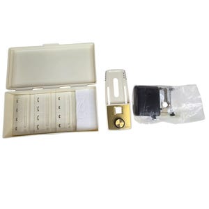 Puede incluir: Una caja de plástico blanquecina abierta que contiene varias piezas pequeñas de plástico beige curvadas. Junto a la caja hay un dispositivo dorado y blanquecino con un botón dorado. Un objeto negro está en una bolsa de plástico transparente.