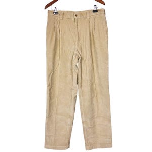 Puede incluir: Pantalones de pana color beige claro colgados de una percha de madera. Los pantalones tienen cierre de botón, trabillas para el cinturón y pliegues en la cintura. La tela de pana tiene un aspecto suave y texturizado. Los pantalones son de color neutro.