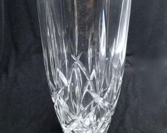 May include: A clear, cut-glass vase with a flared top and a faceted design. The vase features vertical cuts that converge towards the base, creating a diamond-like pattern. The glass is transparent, allowing light to pass through.