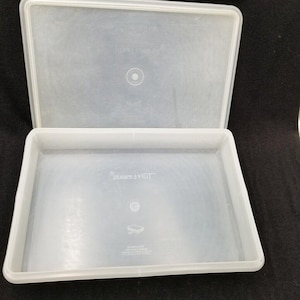 Puede incluir: Un recipiente Tupperware rectangular transparente con una tapa a juego. El recipiente y la tapa están hechos de un material plástico translúcido. La tapa está ligeramente entreabierta, revelando el interior del recipiente. El logotipo de Tupperware es visible.