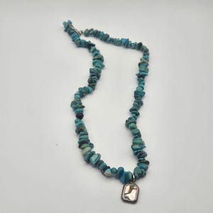 Puede incluir: Un collar compuesto por numerosas piedras irregulares de color turquesa, pulidas y ensartadas, que presenta un único colgante rectangular con un marco metálico y una incrustación de concha iridiscente multicolor en el centro inferior.