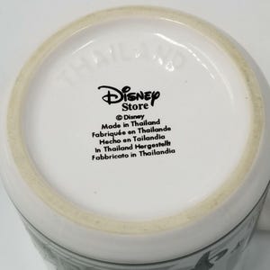 May include: White ceramic mug with the Disney Store logo and text indicating it was made in Thailand. The text is in multiple languages, including English, French, Spanish, German, and Italian. The mug's base is visible.